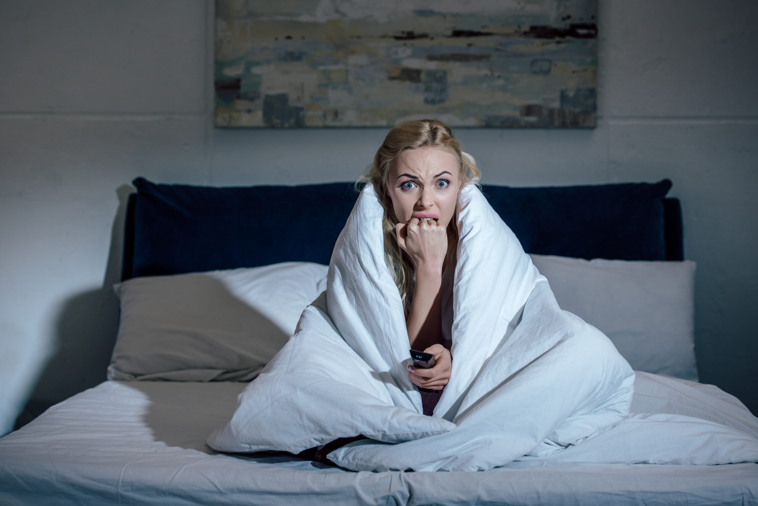 scared woman covered in blanket holding remote control, biting nails and watching tv in bed at home